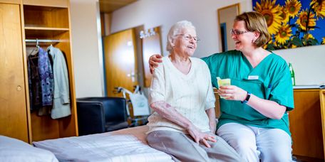 Pflegekraft reicht Seniorin ein Glas Saft,während beide auf dem Pflegebett sitzen