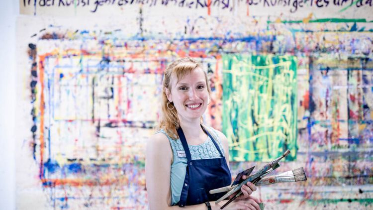 Patientin mit Schürze und Pinseln steht für einer bunten, in der Kunsttherapie gestalteten Wand