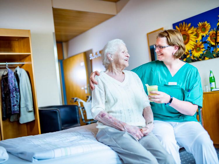 Pflegekraft reicht Seniorin einen Becher mit Saft, während Sie auf dem Bett sitzen