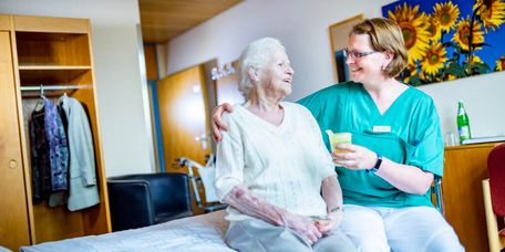 Pflegekraft reicht Seniorin einen Becher mit Saft, während Sie auf dem Bett sitzen
