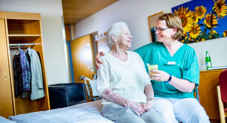 Pflegekraft reicht Seniorin einen Becher mit Saft, während Sie auf dem Bett sitzen