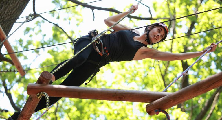 Junger Mann mit Helm klettert im Kletterpark zwischen den Bäumen